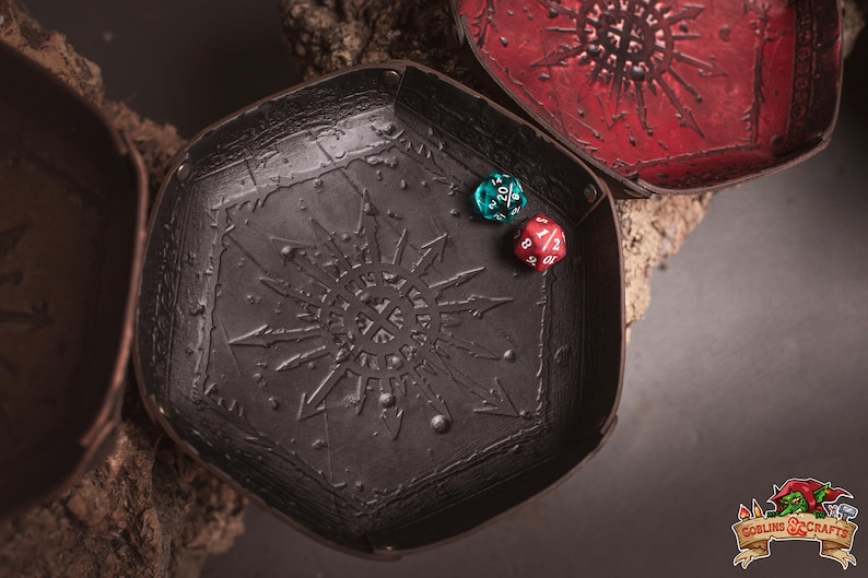May include: A black leather dice tray with a sunburst design and two multi-sided dice. The tray is hexagonal and has a textured surface. A red and a teal die are inside the tray. A red tray is in the background.