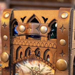 May include: A wooden dice tower with a leather strap and metal accents. The tower is decorated with a gothic design and has a small window on the top. The tower is designed to hold dice and is a great addition to any tabletop game.