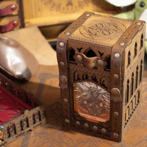 May include: A wooden dice box with a gothic design and a leather strap. The box has a window that shows a miniature figure inside. The box is sitting on a brown surface with a map pattern.