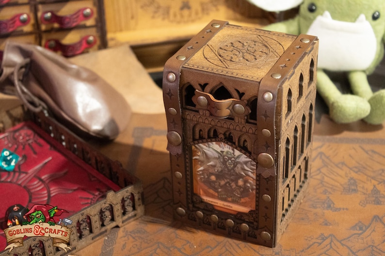 May include: A wooden dice box with a gothic design and a leather strap. The box has a window that shows a miniature figure inside. The box is sitting on a brown surface with a map pattern.