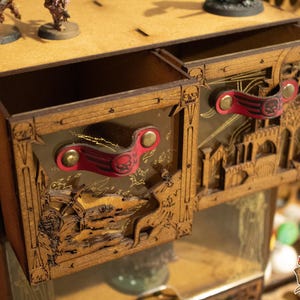 Könnte beinhalten: Miniatur-Malstation aus Holz mit zwei Schubladen. Die Schubladen haben rote Ledergriffe mit Totenkopfmotiven. Die Station ist aufwendig mit einer Landschaft und architektonischen Elementen geschnitzt. Miniaturfiguren befinden sich oben.
