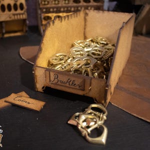 Puede incluir: Una caja de madera con una etiqueta que dice "Buckles" y está llena de hebillas de metal de color dorado. Hay dos etiquetas de madera más pequeñas que dicen "Coins" en la mesa junto a la caja.