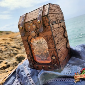 Puede incluir: Cofre de madera con tapa con bisagras, adornado con correas de cuero y tachuelas metálicas. El cofre presenta una imagen enmarcada de una criatura fantástica. El cofre está sobre una tela azul cerca del océano.