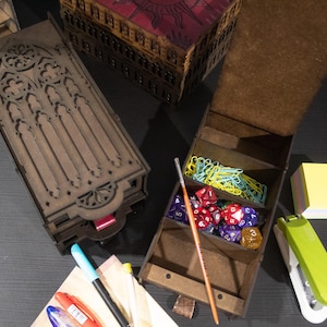 Puede incluir: Dos cajas de madera con diseños intrincados. Una caja tiene un diseño de arco gótico y la otra tiene un compartimento para dados y clips. Las cajas están sobre una superficie negra.