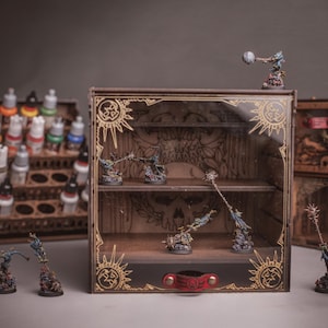 Può includere: Una vetrinetta in legno marrone con pannello frontale trasparente, incisioni dorate a forma di sole sugli angoli e una linguetta in pelle rossa. All'interno, due ripiani ospitano statuette fantasy dipinte, con altre figure e un portacolori accanto.