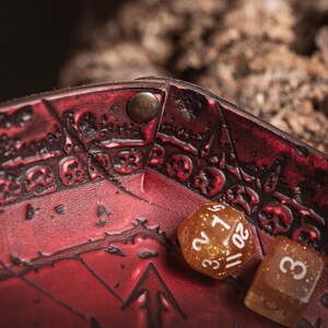 May include: Close-up of a red leather dice tray with embossed skull and arrow designs. Two dice are visible: a gold glitter D20 and a brown D6. The tray is secured with a metal rivet. The Goblin & Crafts logo is in the bottom right corner.