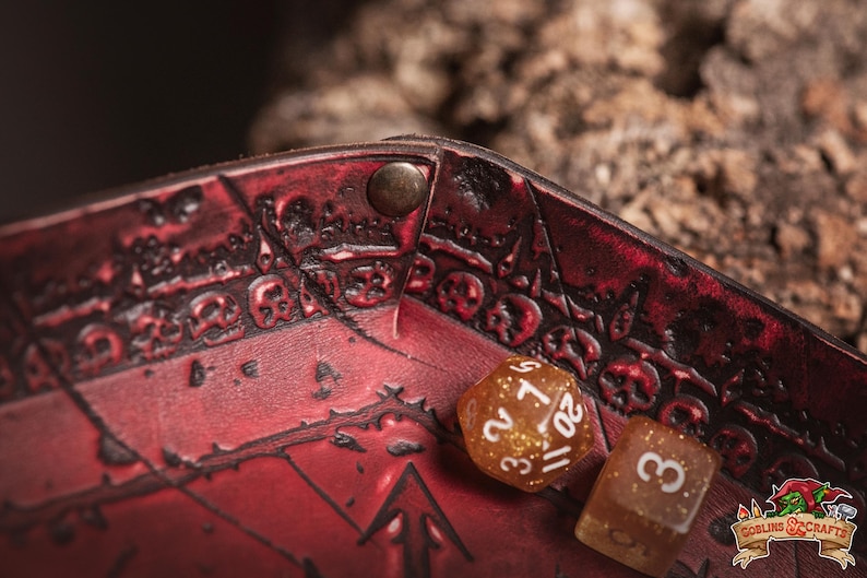 May include: Close-up of a red leather dice tray with embossed skull and arrow designs. Two dice are visible: a gold glitter D20 and a brown D6. The tray is secured with a metal rivet. The Goblin & Crafts logo is in the bottom right corner.