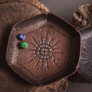 May include: A brown, hexagonal dice tray with a sunburst design and two dice. The tray is made of a textured material and sits on a wooden surface. A second, similar tray is visible.