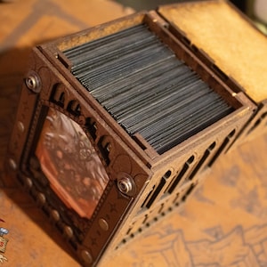 May include: A wooden box with a hinged lid, designed to hold a deck of cards. The box has a decorative design with a small window that shows a picture of a creature. The box is made of dark wood and has metal accents.