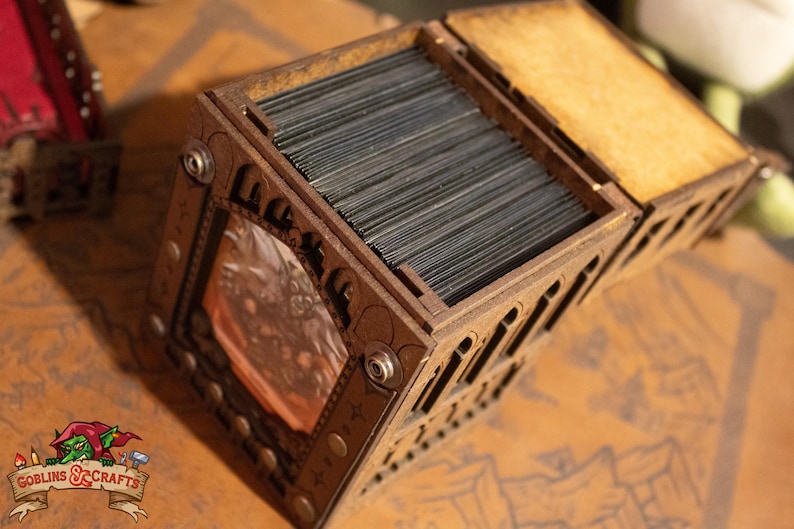 May include: A wooden box with a hinged lid, designed to hold a deck of cards. The box has a decorative design with a small window that shows a picture of a creature. The box is made of dark wood and has metal accents.