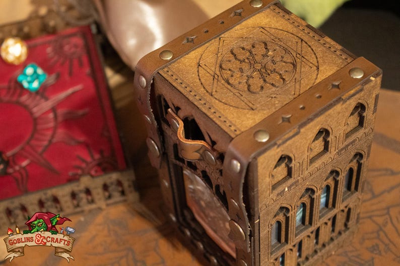 May include: A wooden dice tower with a leather strap and metal accents. The tower is decorated with a carved design and has a small window on the side. The top of the tower has a circular design with a star in the center.
