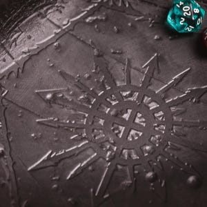 May include: Close-up of a dark gray, textured surface with a sunburst design and two dice. One die is teal with white numbers, and the other is red with white numbers. The image is of a tabletop game accessory.