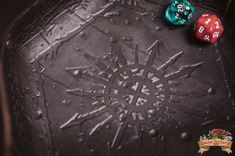 May include: Close-up of a dark gray, textured surface with a sunburst design and two dice. One die is teal with white numbers, and the other is red with white numbers. The image is of a tabletop game accessory.