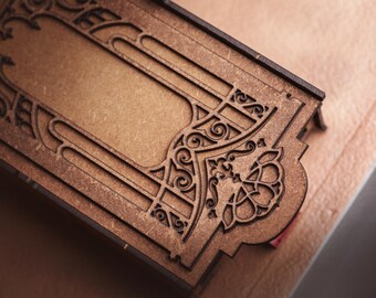 May include: A rectangular wooden box lid featuring intricate laser-cut filigree patterns. The brown wood surface displays a central blank panel framed by symmetrical scrollwork and floral-inspired motifs, showcasing precise cutouts and dark edges.
