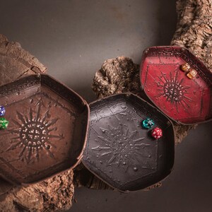 May include: Three hexagonal dice trays in brown, black, and red, each with a sunburst design. Dice of various colors are inside the trays. The trays are displayed on a wooden surface, suitable for tabletop gaming.
