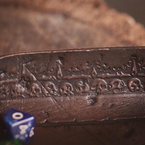 May include: A brown leather dice tray with a detailed embossed skull design. The tray has a riveted edge and a textured surface. A blue and green die are visible in the foreground. The Goblin & Crafts logo is in the bottom right corner.