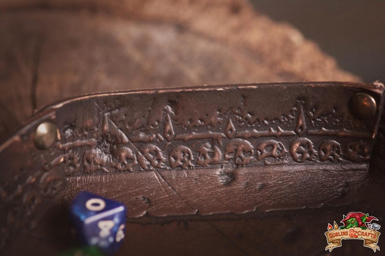 May include: A brown leather dice tray with a detailed embossed skull design. The tray has a riveted edge and a textured surface. A blue and green die are visible in the foreground. The Goblin & Crafts logo is in the bottom right corner.