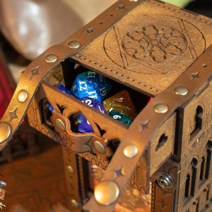 May include: A wooden dice tower with a leather strap and metal accents. The tower is decorated with a carved design and has a hinged lid. The lid is open and there are several dice inside the tower. The dice are blue, green, and purple.