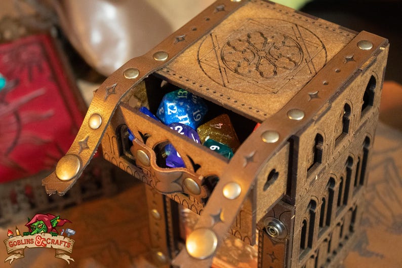 May include: A wooden dice tower with a leather strap and metal accents. The tower is decorated with a carved design and has a hinged lid. The lid is open and there are several dice inside the tower. The dice are blue, green, and purple.