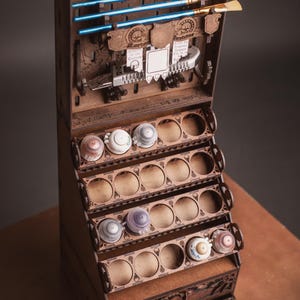 May include: A wooden paint organizer with multiple shelves and drawers. The organizer has slots for paint bottles and brushes. The top section holds blue and tan paint brushes. The organizer is made of brown wood and has decorative cutouts.