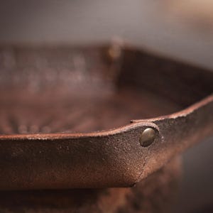 May include: Close-up of a brown leather tray with a riveted corner. The tray has a textured surface and a slightly raised edge. A green and purple die is visible inside the tray. The tray is likely for dice or small items.