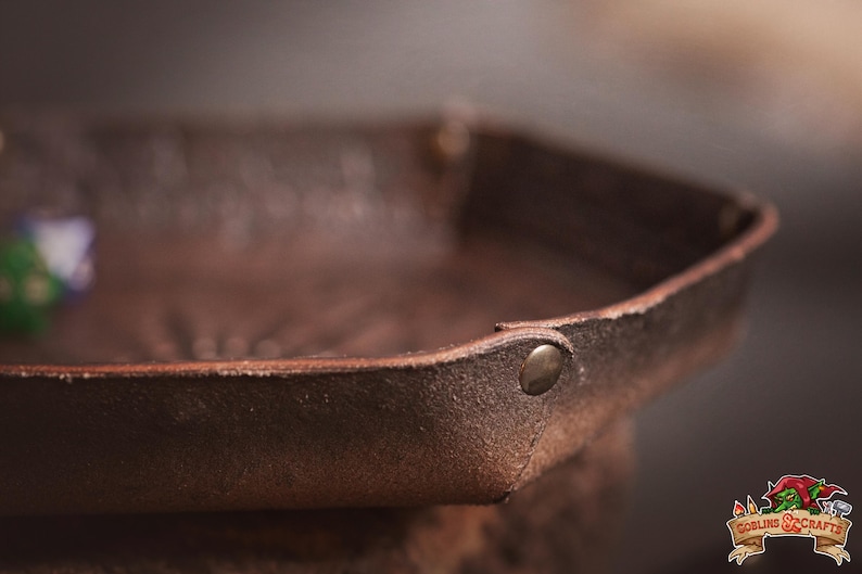 May include: Close-up of a brown leather tray with a riveted corner. The tray has a textured surface and a slightly raised edge. A green and purple die is visible inside the tray. The tray is likely for dice or small items.