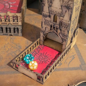 May include: A wooden tower-shaped dice rolling tower with a red felt interior. The tower has a decorative design with a pointed roof and arched windows. There are three dice on the table, one blue, one green, and one gold.