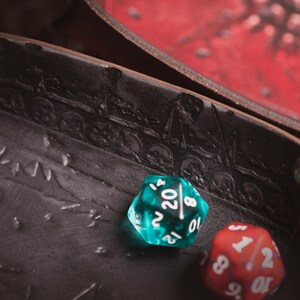 May include: Close-up of a brown leather dice tray with embossed skull and train designs. Two dice are in the tray: a teal D20 and a red D6. The tray is open, revealing a red surface.