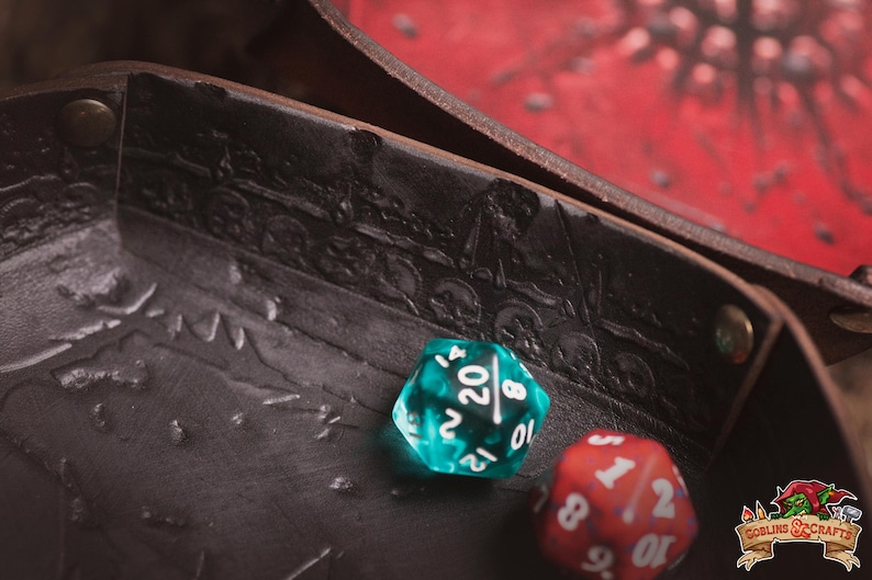 May include: Close-up of a brown leather dice tray with embossed skull and train designs. Two dice are in the tray: a teal D20 and a red D6. The tray is open, revealing a red surface.