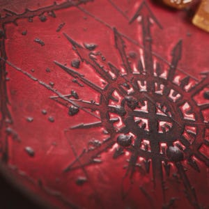 May include: A close-up of a red leather surface with a dark, stylized symbol at the center. The symbol has a star-like design with arrows pointing outwards. The surface has a textured appearance with small, dark accents. A few dice are visible.