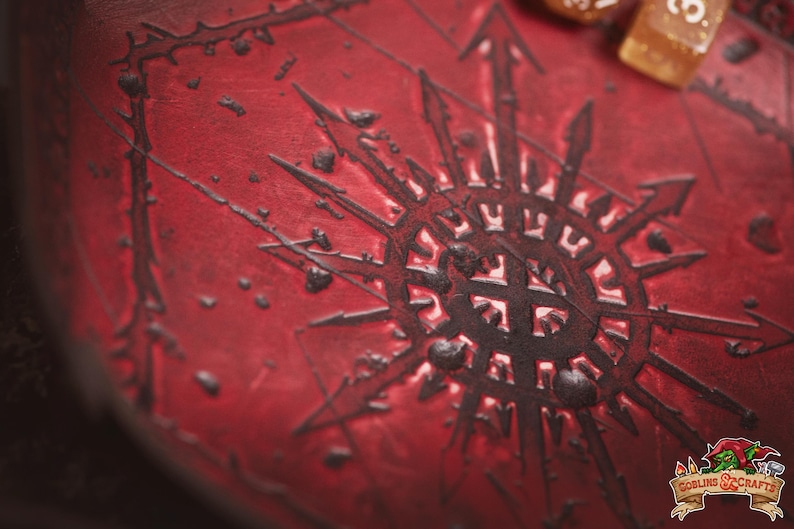May include: A close-up of a red leather surface with a dark, stylized symbol at the center. The symbol has a star-like design with arrows pointing outwards. The surface has a textured appearance with small, dark accents. A few dice are visible.