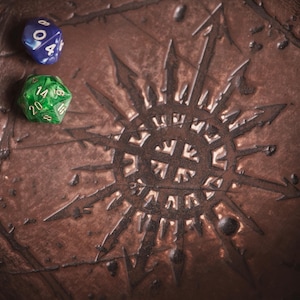 May include: A brown, textured dice tray with a sunburst design and two dice. One die is blue with white numbers, the other is green with white numbers. The tray is likely for tabletop gaming.
