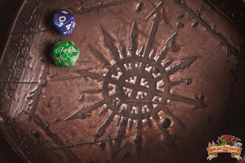 May include: A brown, textured dice tray with a sunburst design and two dice. One die is blue with white numbers, the other is green with white numbers. The tray is likely for tabletop gaming.
