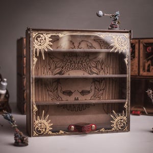 Pode incluir: Uma vitrine quadrada de madeira com três prateleiras horizontais e um painel frontal transparente com desenhos dourados gravados de um sol e uma caveira. A caixa tem um fecho vermelho e está rodeada por pequenas figuras em miniatura.