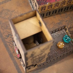 May include: A wooden dice tower with a brown and tan finish. The tower has a decorative design and is open on the top. Two dice are visible inside the tower, one blue and one yellow.