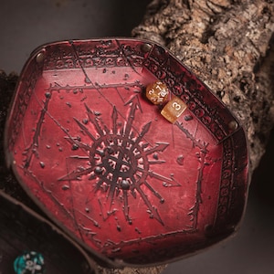 May include: A red, hexagonal dice tray with a detailed, embossed design. The tray features a central sunburst pattern and is made of a textured material. Two dice rest inside the tray. The tray is likely for tabletop gaming.