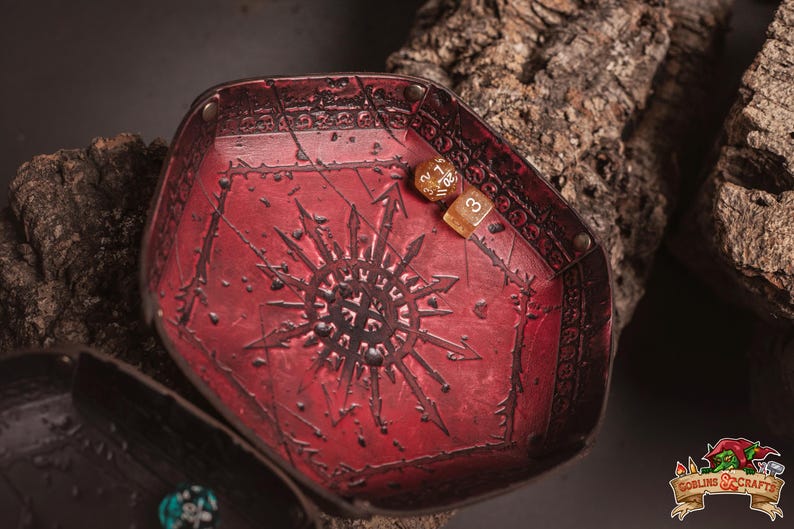 May include: A red, hexagonal dice tray with a detailed, embossed design. The tray features a central sunburst pattern and is made of a textured material. Two dice rest inside the tray. The tray is likely for tabletop gaming.