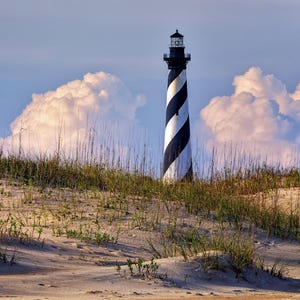 May include: A black and white striped lighthouse stands tall against a backdrop of fluffy white clouds. The lighthouse is situated on a sandy dune covered in green grasses.