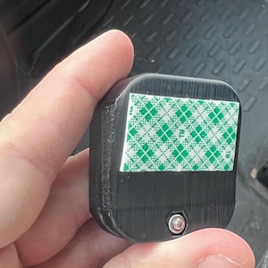 Pode incluir: Um objeto retangular preto com uma superf&iacute;cie texturizada. Uma fita adesiva com padr&atilde;o verde e branco &eacute; aplicada em um lado. Um pequeno parafuso prateado &eacute; vis&iacute;vel na borda inferior do objeto.