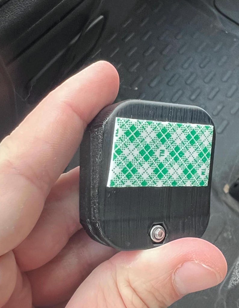 Pode incluir: Um objeto retangular preto com uma superf&iacute;cie texturizada. Uma fita adesiva com padr&atilde;o verde e branco &eacute; aplicada em um lado. Um pequeno parafuso prateado &eacute; vis&iacute;vel na borda inferior do objeto.