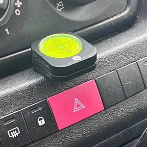 Pode incluir: Uma ferramenta de n&iacute;vel preta com um n&iacute;vel de bolha circular verde n&eacute;on est&aacute; no painel de um ve&iacute;culo. Um bot&atilde;o de luz de emerg&ecirc;ncia rosa brilhante est&aacute; abaixo do n&iacute;vel. Outros bot&otilde;es pretos tamb&eacute;m s&atilde;o vis&iacute;veis.