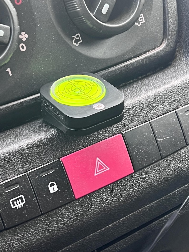 Pode incluir: Uma ferramenta de n&iacute;vel preta com um n&iacute;vel de bolha circular verde n&eacute;on est&aacute; no painel de um ve&iacute;culo. Um bot&atilde;o de luz de emerg&ecirc;ncia rosa brilhante est&aacute; abaixo do n&iacute;vel. Outros bot&otilde;es pretos tamb&eacute;m s&atilde;o vis&iacute;veis.