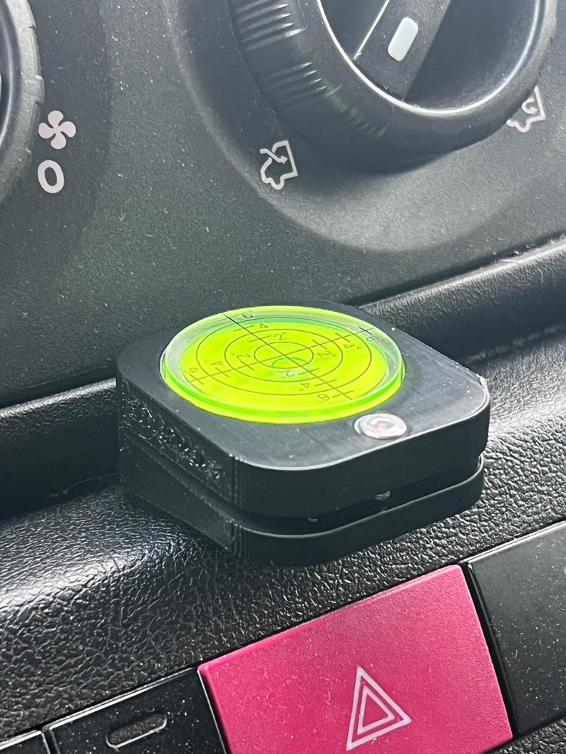 Pode incluir: Um n&iacute;vel preto com um n&iacute;vel de bolha circular verde n&eacute;on. O n&iacute;vel est&aacute; definido em um painel escuro, pr&oacute;ximo a um bot&atilde;o de luz de emerg&ecirc;ncia vermelho. A ferramenta de n&iacute;vel &eacute; quadrada e compacta.