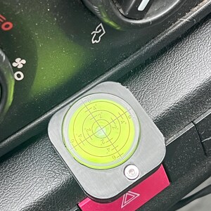 Pode incluir: Uma ferramenta de n&iacute;vel de bolha prateada e amarela com um bot&atilde;o vermelho de luz de emerg&ecirc;ncia. O n&iacute;vel tem um indicador de bolha circular com marca&ccedil;&otilde;es de grau. A ferramenta &eacute; montada em um painel preto.