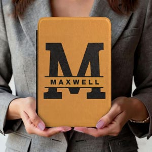 Puede incluir: Una funda de tableta personalizada de color mostaza con una inicial negra "M" y el nombre "MAXWELL" debajo. La funda la sostiene una persona que lleva una blazer gris. El diseño está centrado en la funda, creando un aspecto personalizado y elegante.
