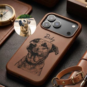 Könnte beinhalten: Eine braune Smartphone-Hülle aus Leder mit einer lasergravierten Illustration eines Hundes und dem darüber gedruckten Namen Rocky. Die Hülle enthält ein kreisförmiges Foto desselben Hundes und einen Ausschnitt für die Dreifachkamera.