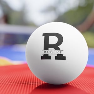 Puede incluir: Una pelota de ping pong blanca con la letra negra "R" y el nombre "ROBERT" impreso debajo. La pelota está sobre una superficie roja. La pelota está diseñada para tenis de mesa.