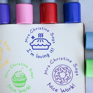 Op de afbeelding: Een verzameling kleurrijke stempels met inktkussens in paars, roze, rood en blauw. De stempels bevatten voedselgerelateerde ontwerpen zoals popcorn, een taart, een cupcake en een donut, met zinnen als "amazing!" en "Nice Work!"