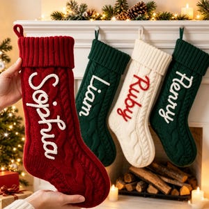 Può includere: Quattro calze natalizie in maglia a trecce con nomi personalizzati in rilievo. Una calza rossa mostra il nome Sophia, mentre altre tre in verde scuro, crema e verde scuro riportano rispettivamente i nomi Liam, Ruby e Henry.