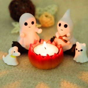 Peut inclure: Une composition décorative sur le thème d'Halloween avec une bougie allumée dans un support rouge et orange, entourée de figurines de fantômes blancs et d'une figurine de petit chien blanc avec un collier jaune. L'ensemble est posé sur une surface claire.